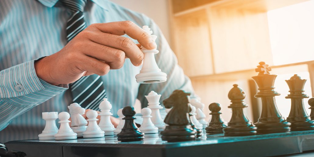 Close up view of a chess board with most all playing pieces on the board. Man wearing a stripped shirt and tie holding white king during a move.