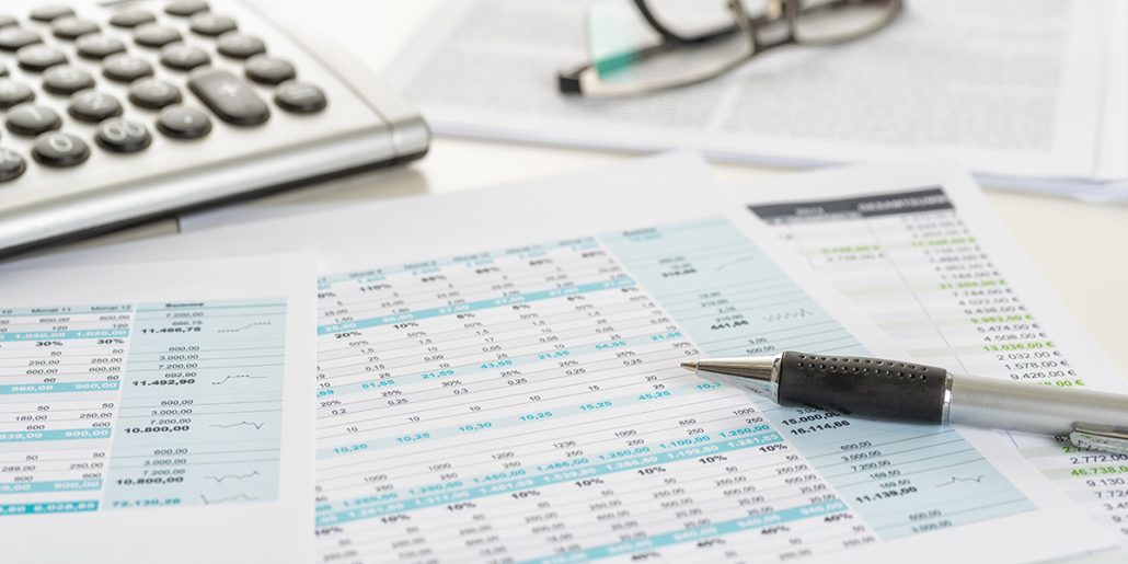 Desktop with accounting sheets, pen, pair of eye glasses and keyboard on desktop.